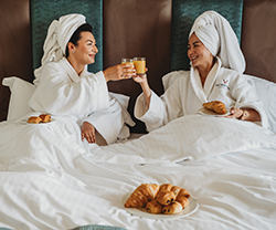 Two ladies in white dressing gowns with hair wrapped in towels sat on a hotel room bed eating pastries and clinking glasses of orange juice